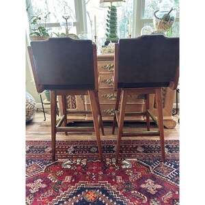 May include: Two brown and wood bar stools with dark brown backs. The stools have a mid-century modern design with a curved back and a footrest. The stools are on a patterned rug.