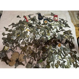 May include: A large collection of vintage keys of various sizes and shapes, scattered across a white surface. Some keys are attached to keychains, including leather fobs and metal tags. The keys are primarily metallic in color.
