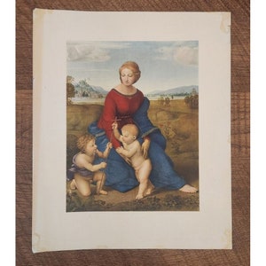 May include: A vintage print of a painting depicting the Madonna and Child with a young Saint John the Baptist. The Madonna is wearing a red and blue robe and is seated on a grassy hill. The Christ Child is standing and holding a cross, while Saint John the Baptist is kneeling and holding a reed.