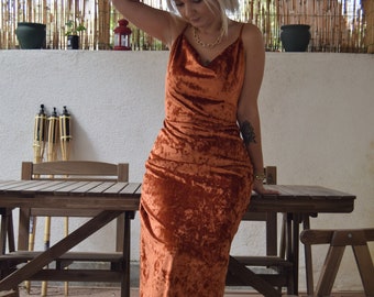 Könnte beinhalten: Ein rostfarbenes Maxikleid aus Samt mit Spaghettiträgern und einem Wasserfallausschnitt. Das Kleid ist bodenlang und hat ein drapiertes, gerafftes Design. Das Model steht in der Nähe eines Holztisches und Stühlen.
