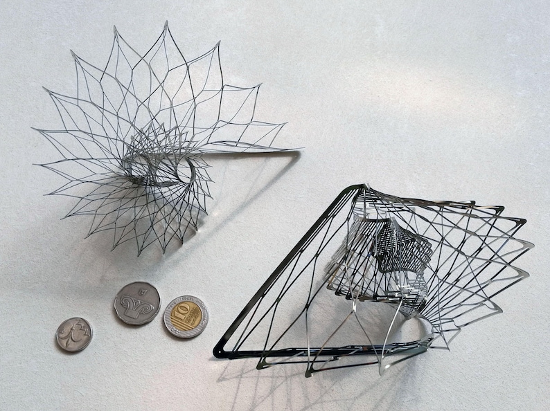 May include: Two silver wire sculptures, one resembling a spiral and the other a geometric shape, are displayed on a white surface. Three silver coins are also visible.
