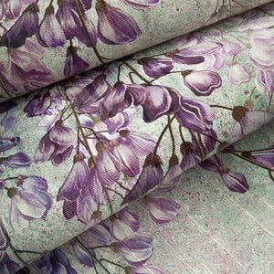 May include: A textile fabric featuring a repeating pattern of purple wisteria flowers and buds set against a light mint green background. The material displays a textured weave with visible vertical lines and a subtle speckled effect throughout.