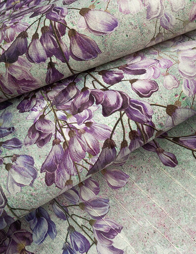 May include: A textile fabric featuring a repeating pattern of purple wisteria flowers and buds set against a light mint green background. The material displays a textured weave with visible vertical lines and a subtle speckled effect throughout.