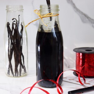 May include: A clear glass bottle filled with vanilla beans, a dark brown glass bottle filled with vanilla extract, and a spool of red glitter ribbon.