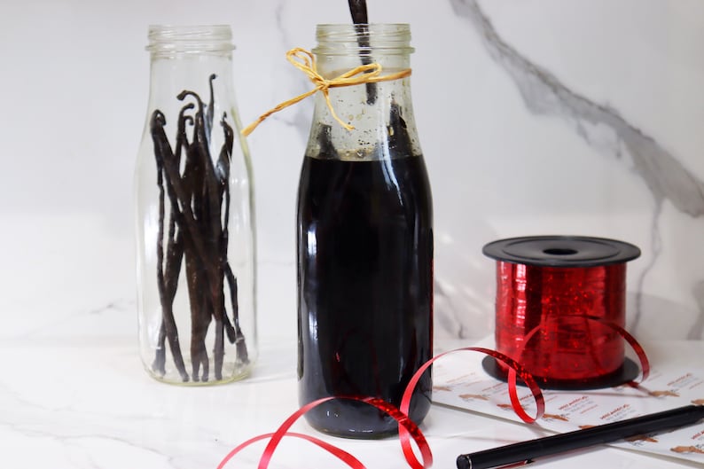 May include: A clear glass bottle filled with vanilla beans, a dark brown glass bottle filled with vanilla extract, and a spool of red glitter ribbon.