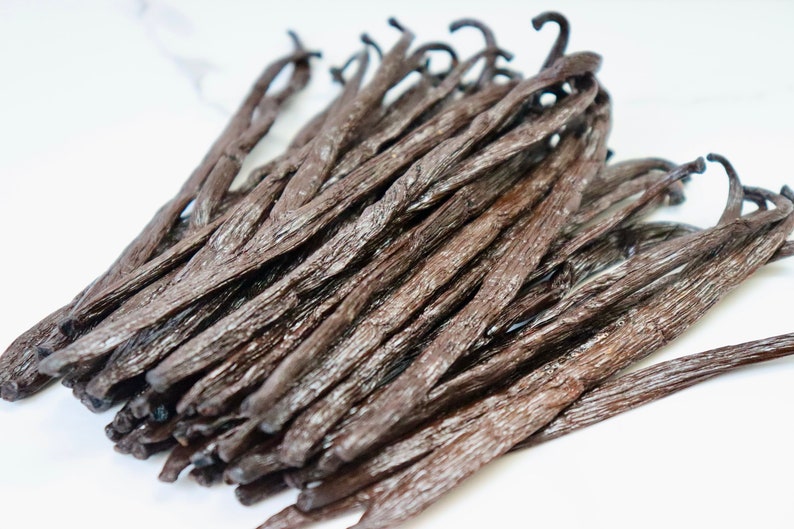 May include: A close-up of a pile of dried vanilla beans. The beans are dark brown and have a shiny, slightly oily surface. They are arranged in a random pattern, with some beans overlapping others.