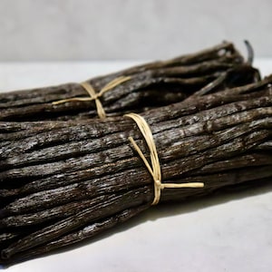 Puede incluir: Dos paquetes de vainas de vainilla secas atadas con cuerda. Las vainas son de color marrón oscuro y tienen un acabado brillante.