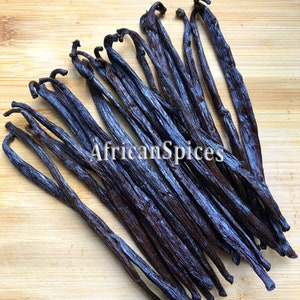 May include: A bunch of dark brown vanilla beans on a light brown wooden surface. The beans are arranged in a fan shape, with the tips of the beans pointing towards the top of the image. The text "AfricanSpices" is visible in the center of the image.