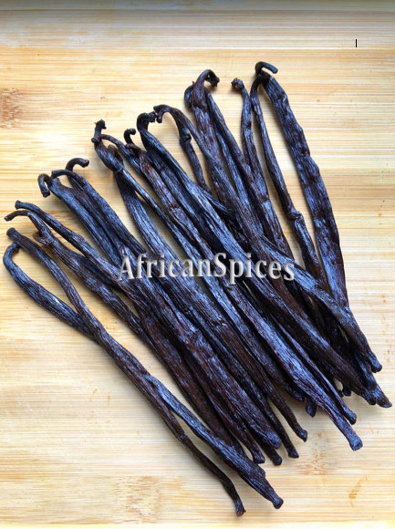 May include: A bunch of dark brown vanilla beans on a light brown wooden surface. The beans are arranged in a fan shape, with the tips of the beans pointing towards the top of the image. The text "AfricanSpices" is visible in the center of the image.
