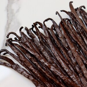 May include: A close-up of a bunch of dark brown vanilla beans on a white marble surface.