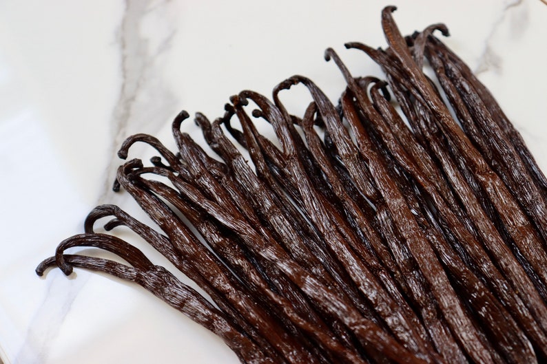 May include: A close-up of a bunch of dark brown vanilla beans on a white marble surface.