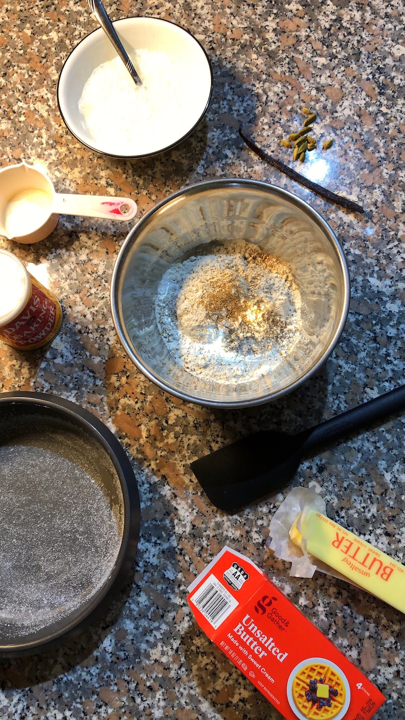 May include: A close-up of a kitchen counter with baking ingredients, including flour, sugar, butter, and vanilla beans. There is also a baking pan and a bowl of cream.