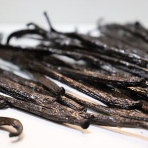 May include: A close-up of a group of dried vanilla beans. The beans are dark brown and have a shiny, slightly oily surface. They are arranged in a random pattern, with some beans overlapping others.