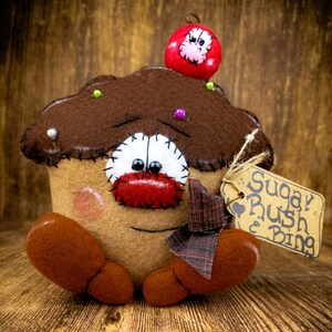 May include: A brown felt cupcake with a red felt cherry on top. The cupcake has a stitched-on face with googly eyes and a red felt nose. The cupcake is sitting on a wooden surface and has a tag that says "Sugary Rush & Bing".