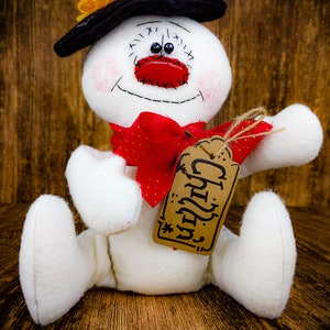 Puede incluir: Un mu&ntilde;eco de nieve blanco con un sombrero negro, un lazo rojo y una etiqueta marr&oacute;n que dice "Chilly".