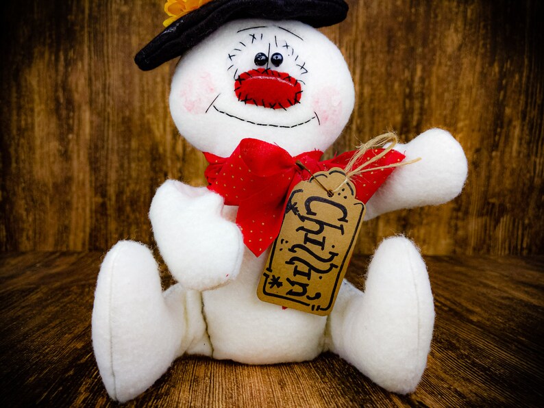 Puede incluir: Un mu&ntilde;eco de nieve blanco con un sombrero negro, un lazo rojo y una etiqueta marr&oacute;n que dice "Chilly".