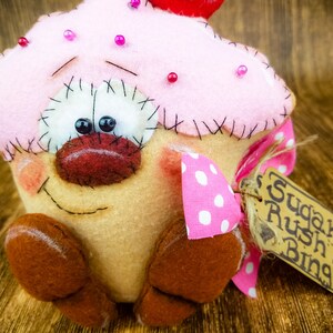 May include: A pink and brown felt cupcake with a red cherry on top. The cupcake has a smiling face with black eyes and a red nose. It is wearing a pink and white polka dot bow and has a brown tag that says "Sugar Rush Bing".