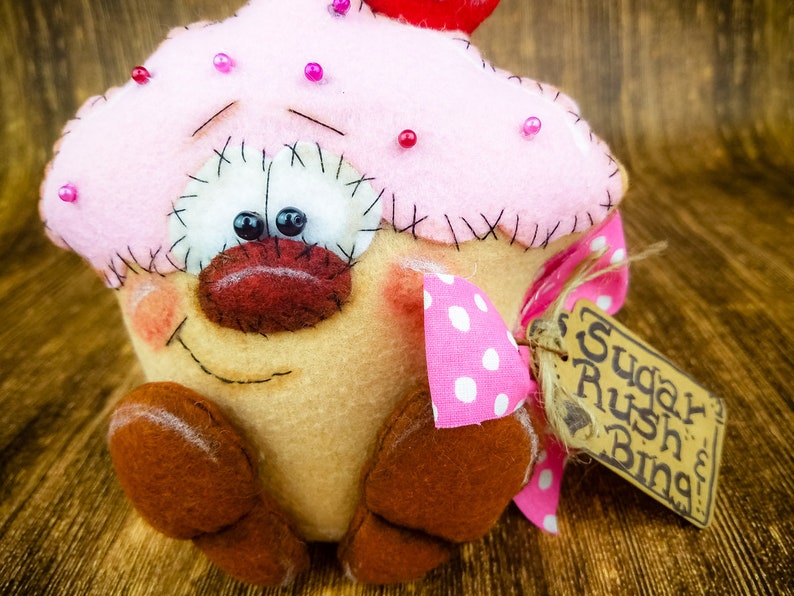 May include: A pink and brown felt cupcake with a red cherry on top. The cupcake has a smiling face with black eyes and a red nose. It is wearing a pink and white polka dot bow and has a brown tag that says "Sugar Rush Bing".