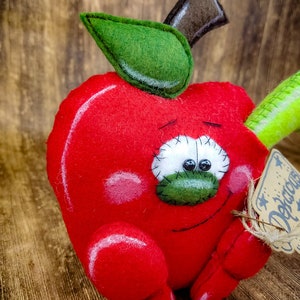 May include: A red felt apple with a green felt leaf and stem. The apple has a stitched-on face with googly eyes and a stitched-on smile. The apple is holding a green felt pencil in its right hand and a brown tag with the word "Delicious" written on it in white.
