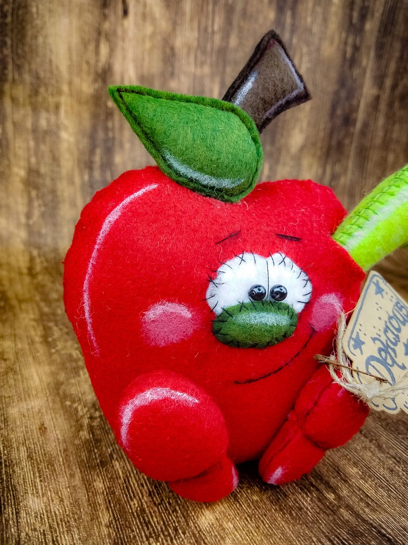 May include: A red felt apple with a green felt leaf and stem. The apple has a stitched-on face with googly eyes and a stitched-on smile. The apple is holding a green felt pencil in its right hand and a brown tag with the word "Delicious" written on it in white.