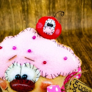 May include: A pink felt cupcake with a red felt cherry on top. The cupcake has a stitched face with black eyes and a red mouth. The cherry has black eyes and a stitched smile. The cupcake is decorated with pink beads.