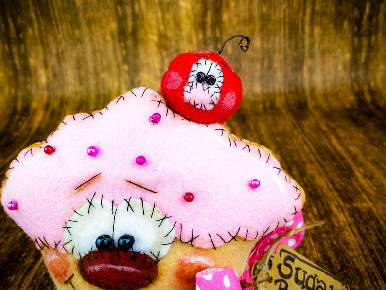 May include: A pink felt cupcake with a red felt cherry on top. The cupcake has a stitched face with black eyes and a red mouth. The cherry has black eyes and a stitched smile. The cupcake is decorated with pink beads.