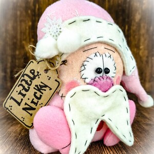 May include: A plush doll featuring a pink hat with a white snowflake and pom-poms, a peach-coloured face with black stitched eyes, a pink nose, and a white beard. A brown cardboard tag attached with twine displays the text Little Nicky.