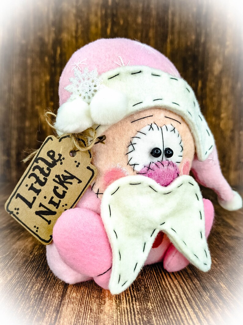May include: A plush doll featuring a pink hat with a white snowflake and pom-poms, a peach-coloured face with black stitched eyes, a pink nose, and a white beard. A brown cardboard tag attached with twine displays the text Little Nicky.