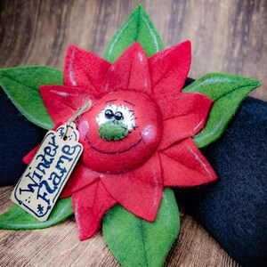 May include: A handmade felt poinsettia flower with a smiling face and green leaves. The red flower has a tag that reads "Winter Flame". The poinsettia is set against a dark background and a wooden surface.