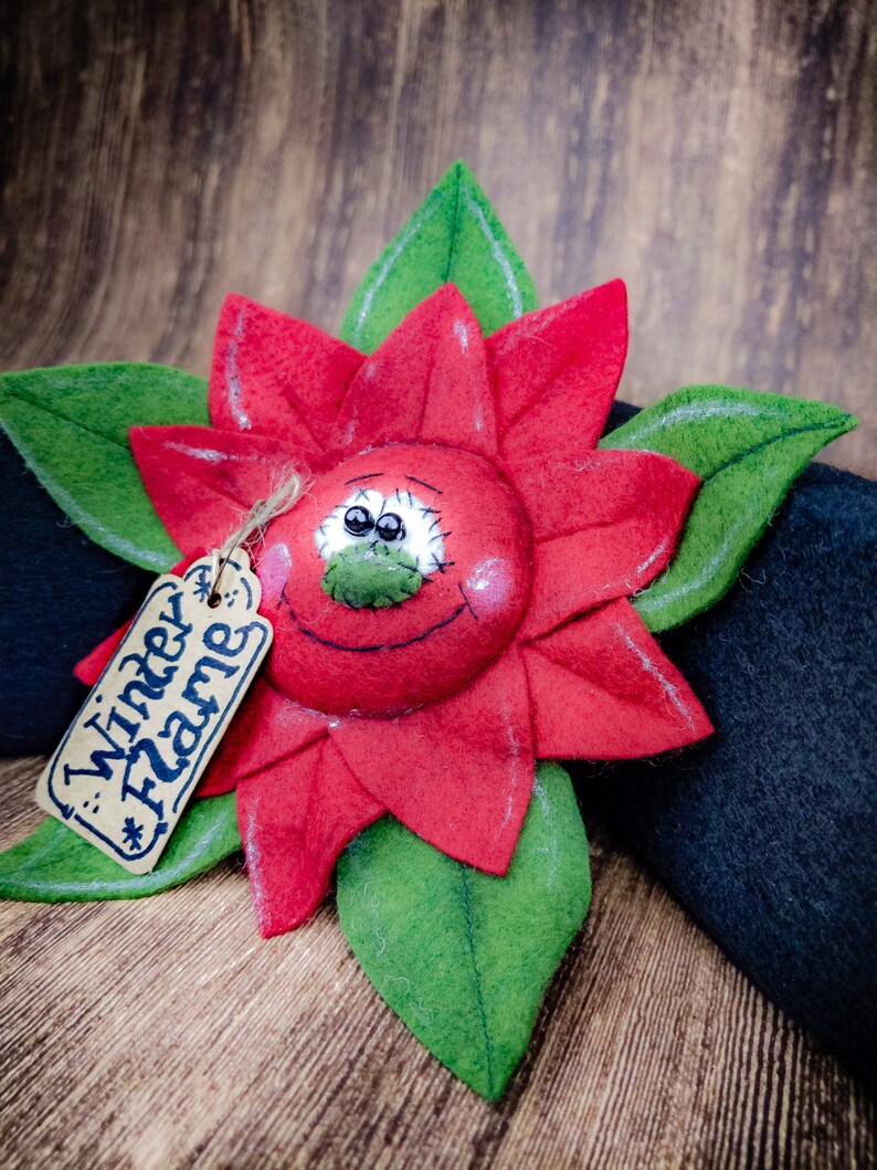 May include: A handmade felt poinsettia flower with a smiling face and green leaves. The red flower has a tag that reads "Winter Flame". The poinsettia is set against a dark background and a wooden surface.
