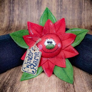 May include: A handcrafted poinsettia flower decoration with red petals and green leaves. The flower has a smiling face and a tag that reads "Winter Flame". The decoration is placed on a dark surface against a wooden background.