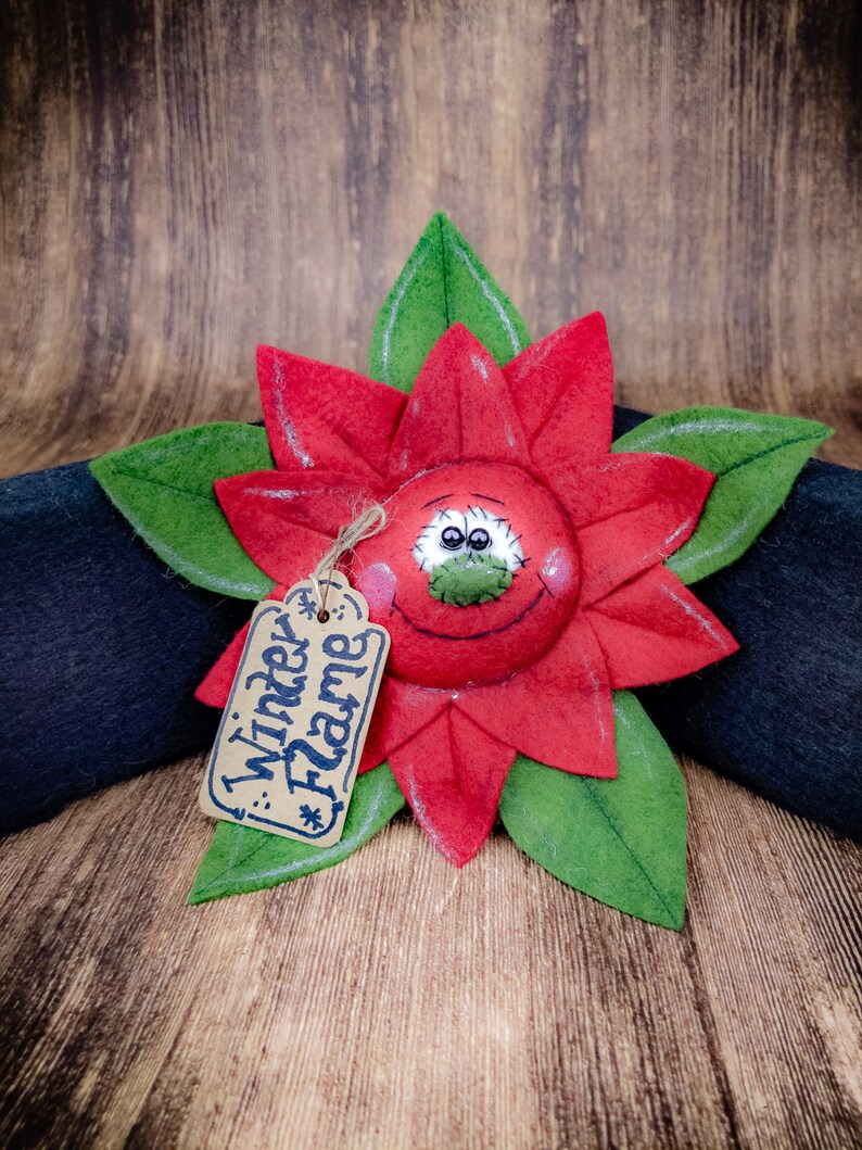 May include: A handcrafted poinsettia flower decoration with red petals and green leaves. The flower has a smiling face and a tag that reads "Winter Flame". The decoration is placed on a dark surface against a wooden background.