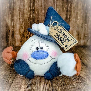 May include: A white felt snowman with a blue felt hat, blue felt eyes, and a blue felt mouth. The snowman is holding a white felt snowball in one hand and has a brown felt mitten on the other hand. The snowman has a brown felt tag that says "Snow Ball" hanging from its hat.