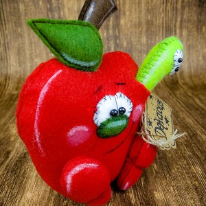 May include: A red felt apple with a green felt leaf and a brown felt stem. The apple has a stitched-on face with black eyes and a green felt mouth. A green felt worm is attached to the side of the apple. The apple has a brown tag with the word "Decor" written on it.