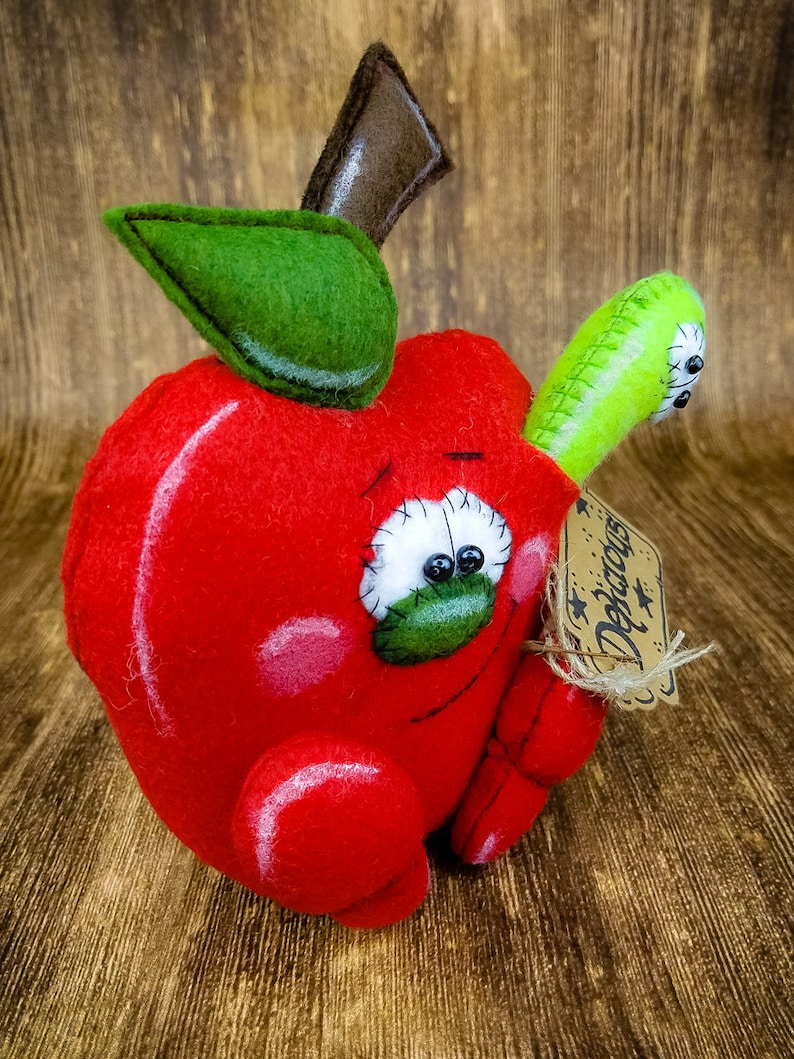 May include: A red felt apple with a green felt leaf and a brown felt stem. The apple has a stitched-on face with black eyes and a green felt mouth. A green felt worm is attached to the side of the apple. The apple has a brown tag with the word "Decor" written on it.
