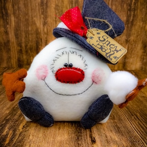 May include: A white snowman-shaped plush toy with a red nose, black eyes, and a black stitched smile. The snowman is wearing a blue hat with a red bow and a brown tag that says "Snow Ball".