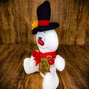 Puede incluir: Un mu&ntilde;eco de nieve blanco con un sombrero de copa negro, un lazo rojo y una etiqueta marr&oacute;n que dice "Chilly". El mu&ntilde;eco de nieve est&aacute; sentado sobre una superficie de madera.