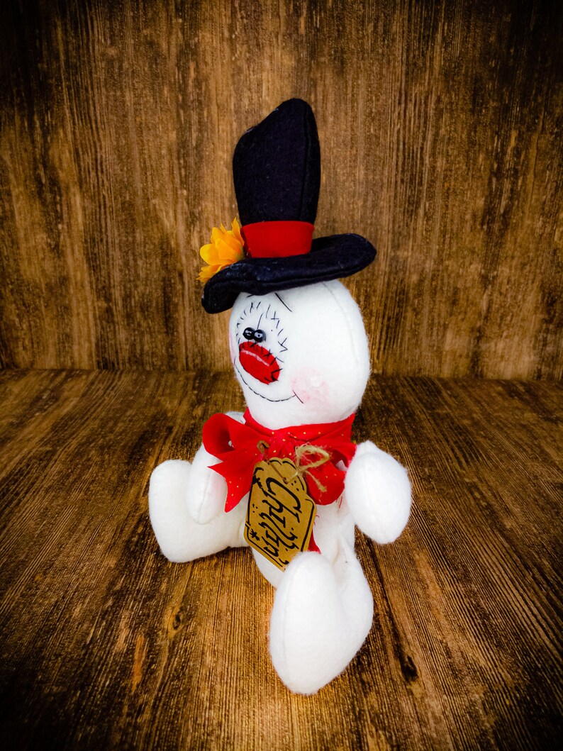 Puede incluir: Un mu&ntilde;eco de nieve blanco con un sombrero de copa negro, un lazo rojo y una etiqueta marr&oacute;n que dice "Chilly". El mu&ntilde;eco de nieve est&aacute; sentado sobre una superficie de madera.