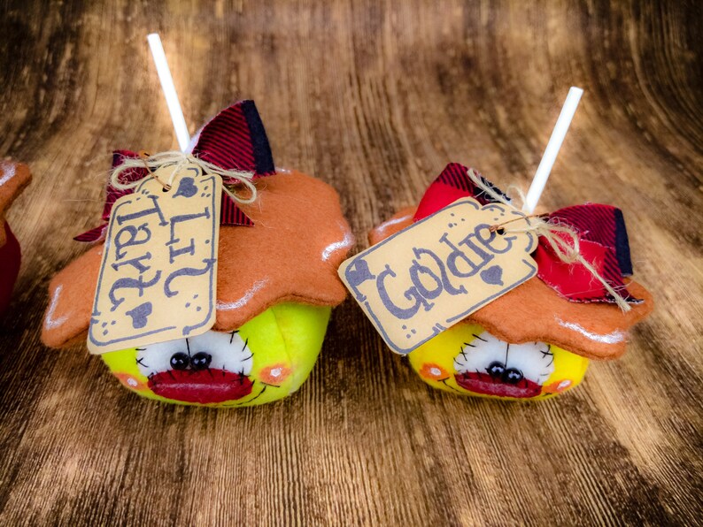 May include: Two handmade felt apple decorations with brown felt tops and yellow felt bottoms. The apples have black eyes, red mouths, and brown felt stems. Each apple has a brown paper tag tied with twine. The tags read "Lil Tart" and "Goldie".