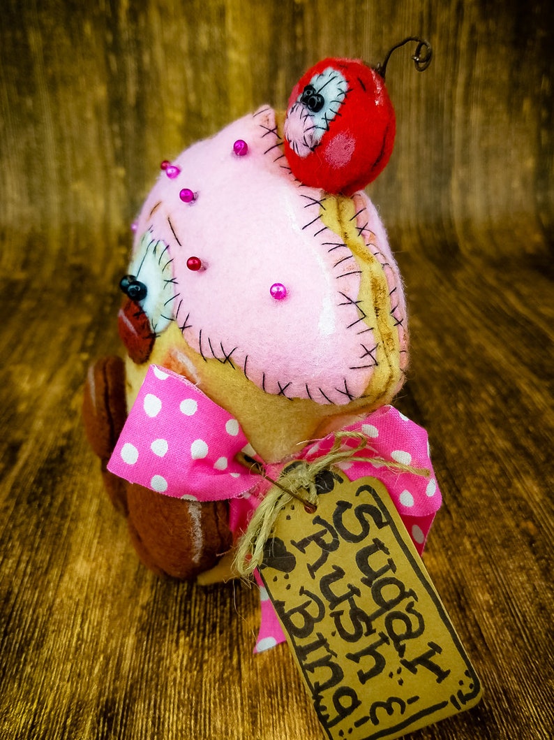 May include: A pink and brown felt cupcake with a red felt cherry on top. The cupcake has pink polka dots and a brown felt base. It has a tag that says "Happy Birthday" with a heart and the words "Rush Bind".