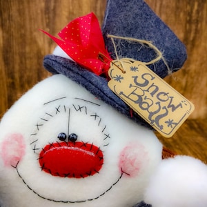 May include: A white snowman with a red nose and black eyes, wearing a blue hat with a red bow and a brown tag that says "Snow Ball".