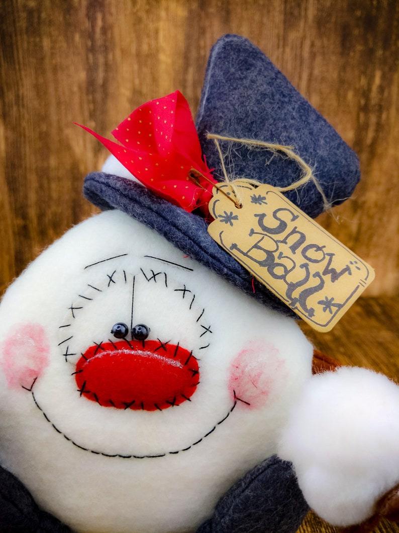May include: A white snowman with a red nose and black eyes, wearing a blue hat with a red bow and a brown tag that says "Snow Ball".