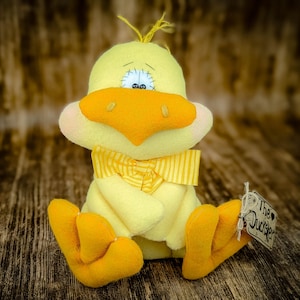May include: A yellow plush duck with a yellow and white striped bow tie. The duck has large, round eyes and a small, orange beak. The duck is sitting on a wooden surface with a brown tag that says "The Duckie" hanging from its foot.