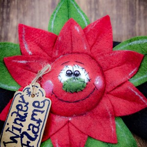 May include: A handcrafted poinsettia flower decoration with a smiling face and the text "Winter Flame" on a tag. The flower has red petals, green leaves, and a black base, perfect for holiday decor.