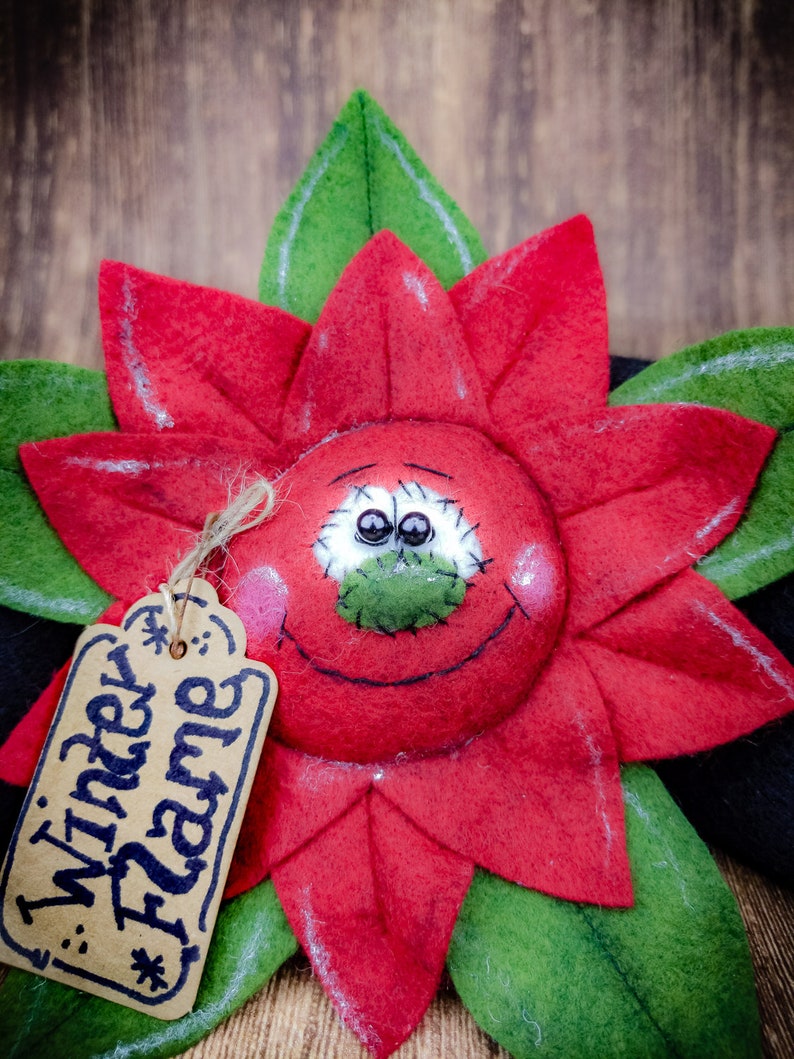 May include: A handcrafted poinsettia flower decoration with a smiling face and the text "Winter Flame" on a tag. The flower has red petals, green leaves, and a black base, perfect for holiday decor.