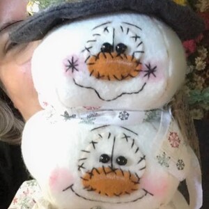 Pu&ograve; includere: Una pila di tre decorazioni di pupazzo di neve bianche con occhi neri e una bocca di feltro marrone. Il pupazzo di neve in cima indossa un cappello a cilindro nero con una fascia rossa e un pon pon bianco.