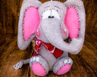 Puede incluir: Un juguete de elefante de fieltro gris con orejas y pies rosas. El elefante lleva una bufanda de lunares rojos y blancos. El juguete está sentado sobre una superficie de madera.