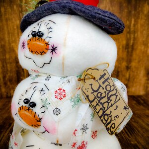 Pu&ograve; includere: Un pupazzo di neve bianco con un cappello nero e una sciarpa a motivi rossi e bianchi. Il pupazzo di neve ha un'etichetta marrone che dice "Buone Feste".