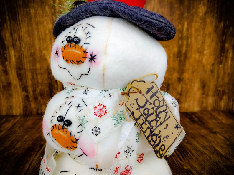Pu&ograve; includere: Un pupazzo di neve bianco con un cappello nero e una sciarpa a motivi rossi e bianchi. Il pupazzo di neve ha un'etichetta marrone che dice "Buone Feste".