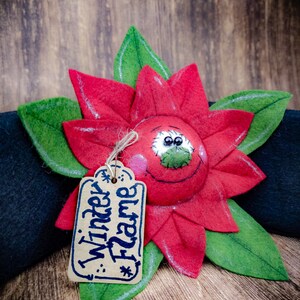 May include: A handmade felt poinsettia flower with green leaves and a smiling face. A small tag with the words "Winter Flame" is attached. The flower is red with white stitching and pink cheeks.